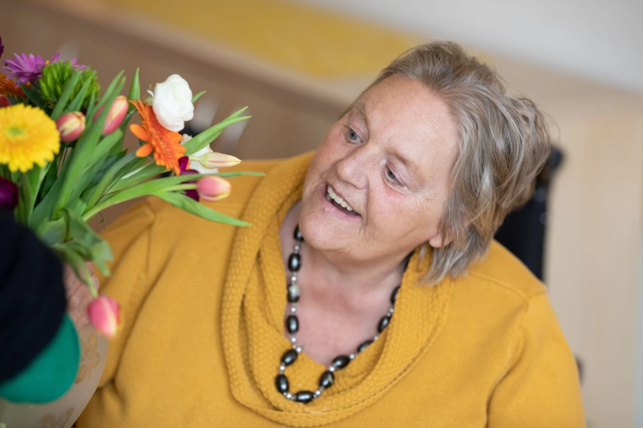 Eine Frau in gelbem Pullover l&auml;chelt und schaut auf einen bunten Blumenstrau&szlig;. Sie tr&auml;gt eine auff&auml;llige Perlenkette.