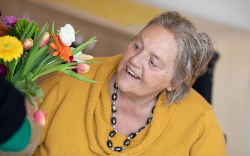 Eine Frau in gelbem Pullover l&auml;chelt und schaut auf einen bunten Blumenstrau&szlig;. Sie tr&auml;gt eine auff&auml;llige Perlenkette.