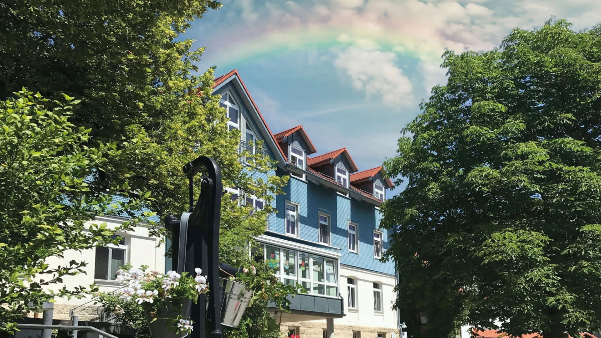 Blau-wei&szlig;es historisch ausstehendes Geb&auml;ude umgeben von gr&uuml;nen B&auml;umen und Blumenbeet. Am Himmel steht ein Regenbogen.