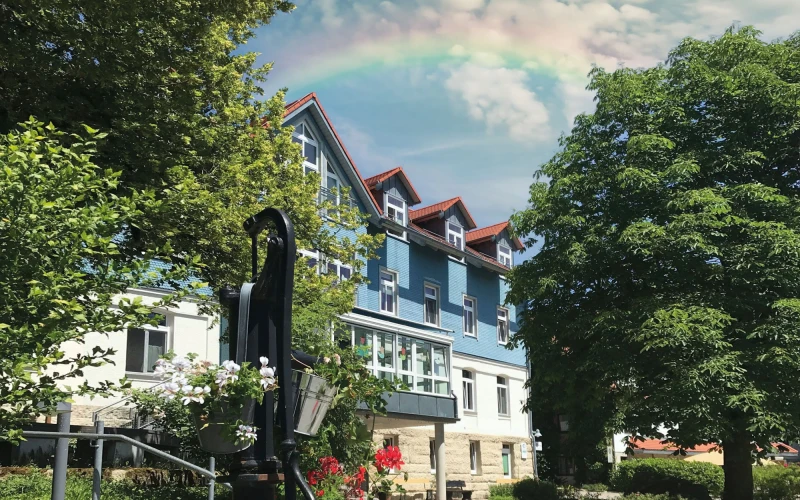 Blau-wei&szlig;es historisch ausstehendes Geb&auml;ude umgeben von gr&uuml;nen B&auml;umen und Blumenbeet. Am Himmel steht ein Regenbogen.