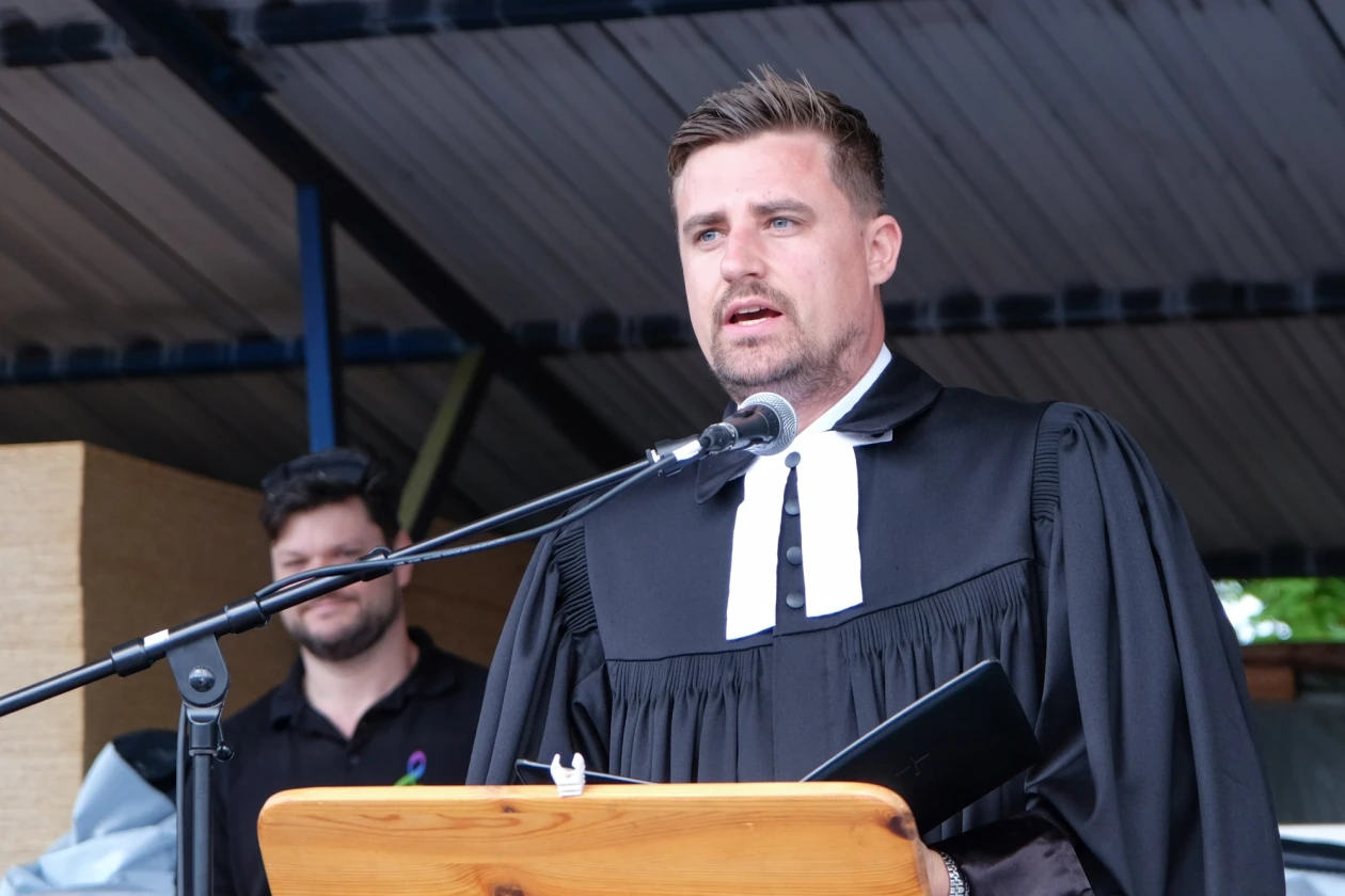 Ein Mann im Talar steht vorne am Rednerpult und h&auml;lt ein ge&ouml;ffnetes Buch in der Hand. Im Hintergrund ist eine weitere Person zu sehen.