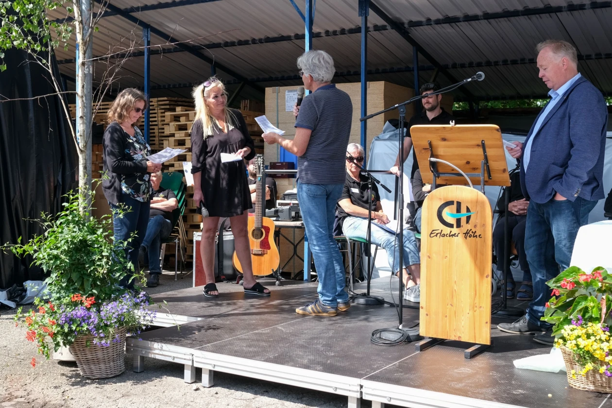 Menschen stehen auf einer B&uuml;hne. Ein Mann mit grauen Haaren spricht mit einer blondhaarigen Frau in schwarzem Kleid. Ein Rednerpult mit Aufschrift "Erlacher H&ouml;he steht im Vordergrund. Blumen schm&uuml;cken die B&uuml;hne.