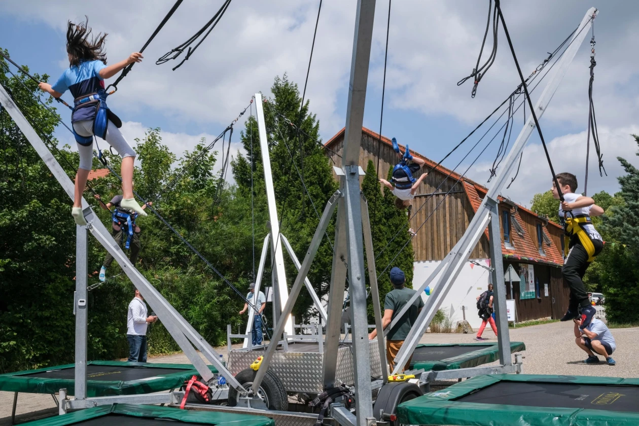 Ein Bungee-Trampolin ist aufgebaut. Kinder h&uuml;pfen auf vier Seiten des Sportger&auml;tes, w&auml;hrend Menschen im Hintergrund zuschauen.