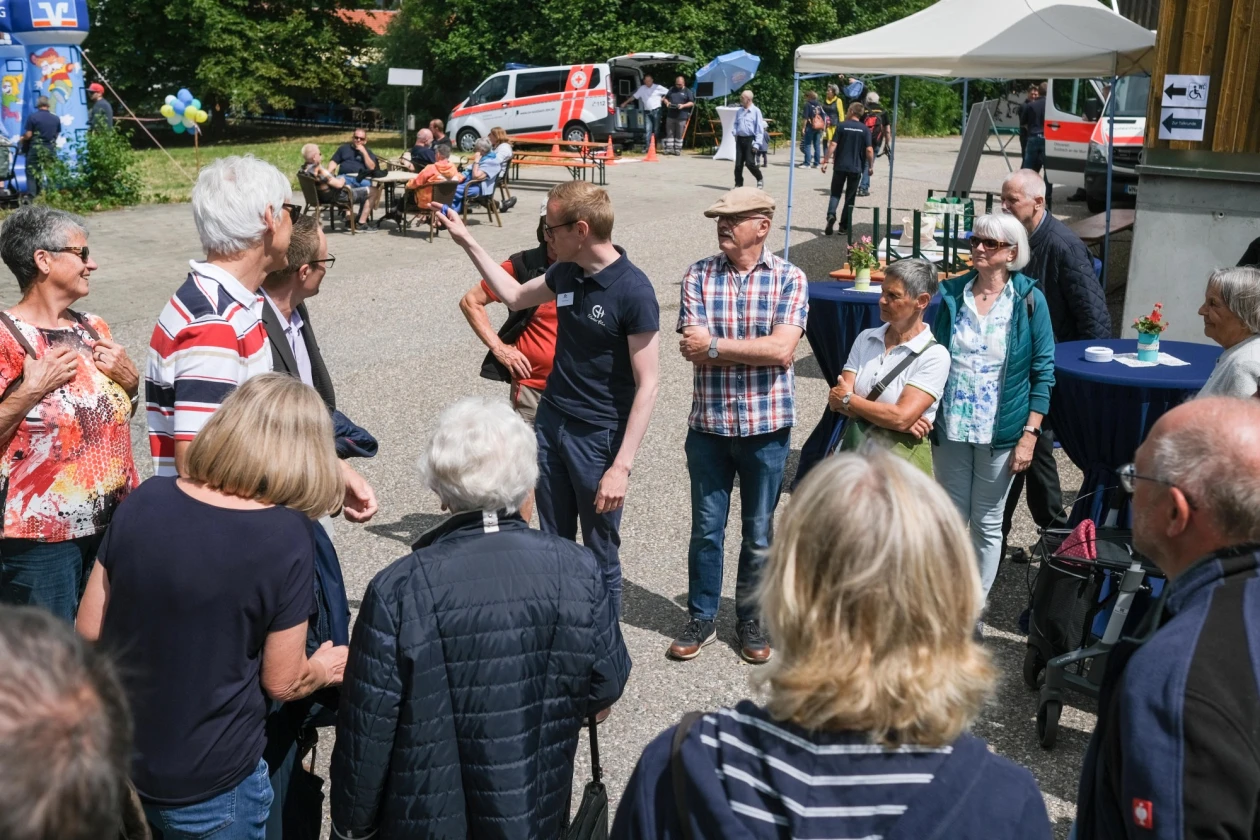 Eine Menschengruppe steht um einen jungen Mann mit dunklem T-Shirt und Logo der "Erlacher H&ouml;he". Im Hintergrund ist umtriebiger Festbetrieb zu sehen.