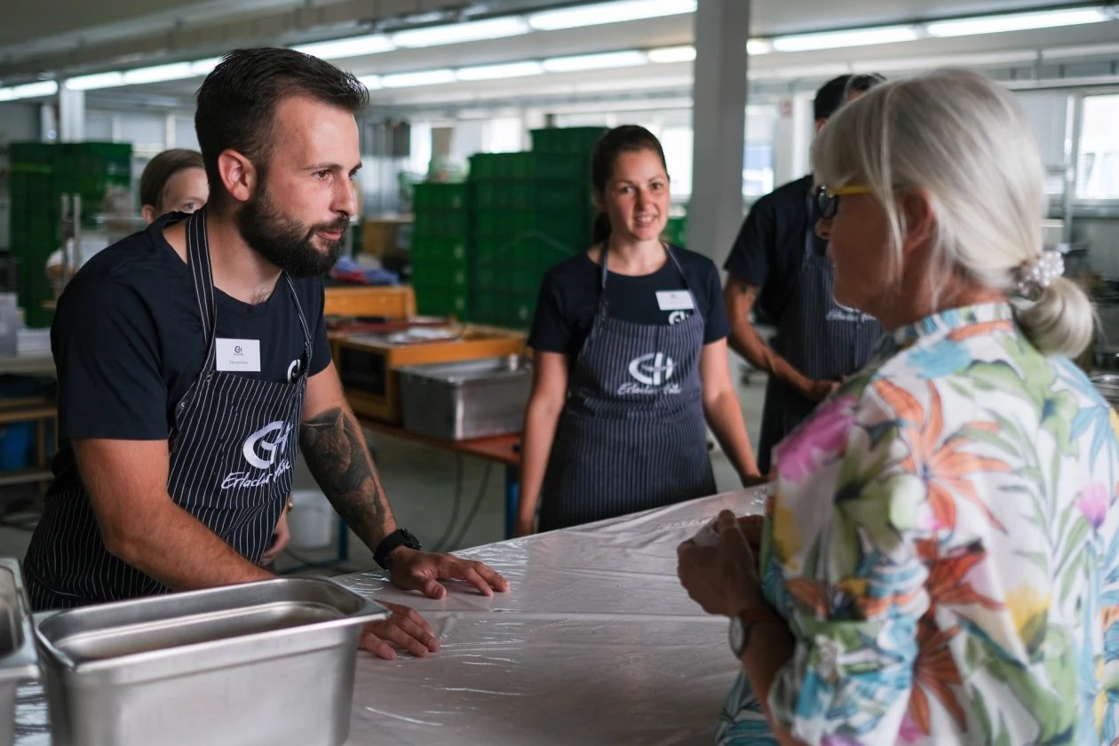 An einer Theke stehen vier Menschen in dunklem T-Shirt und dunkler Sch&uuml;rze mit dem Logo der "Erlacher H&ouml;he". Im Vordergrund steht eine weitere Frau in einem Oberteil mit Blumenmuster.