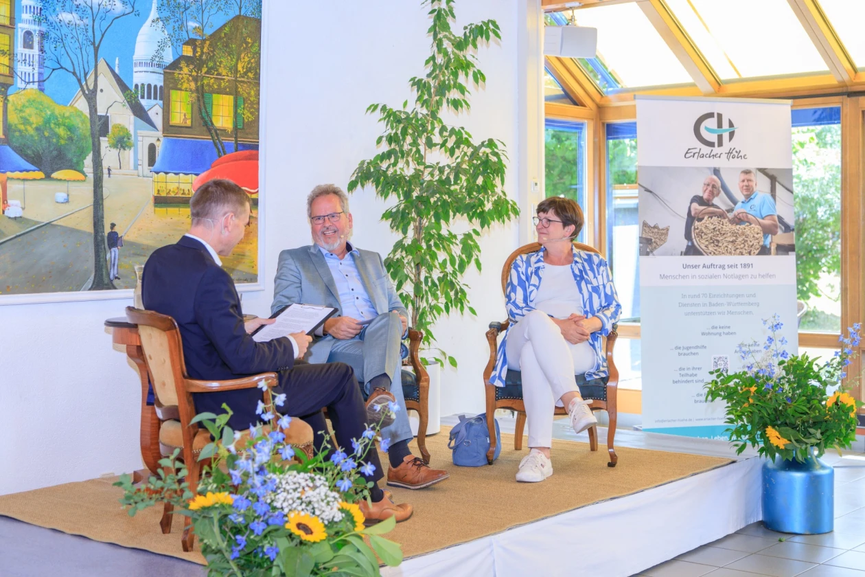 Drei Personen sitzen auf einer B&uuml;hne. Der Mann links h&auml;lt ein Dokument in den H&auml;nden. Im Vordergrund stehen zwei Blumenstr&auml;u&szlig;e. Im Hintergrund ist ein gro&szlig;es Wandbild und ein Roll-up zu sehen.
