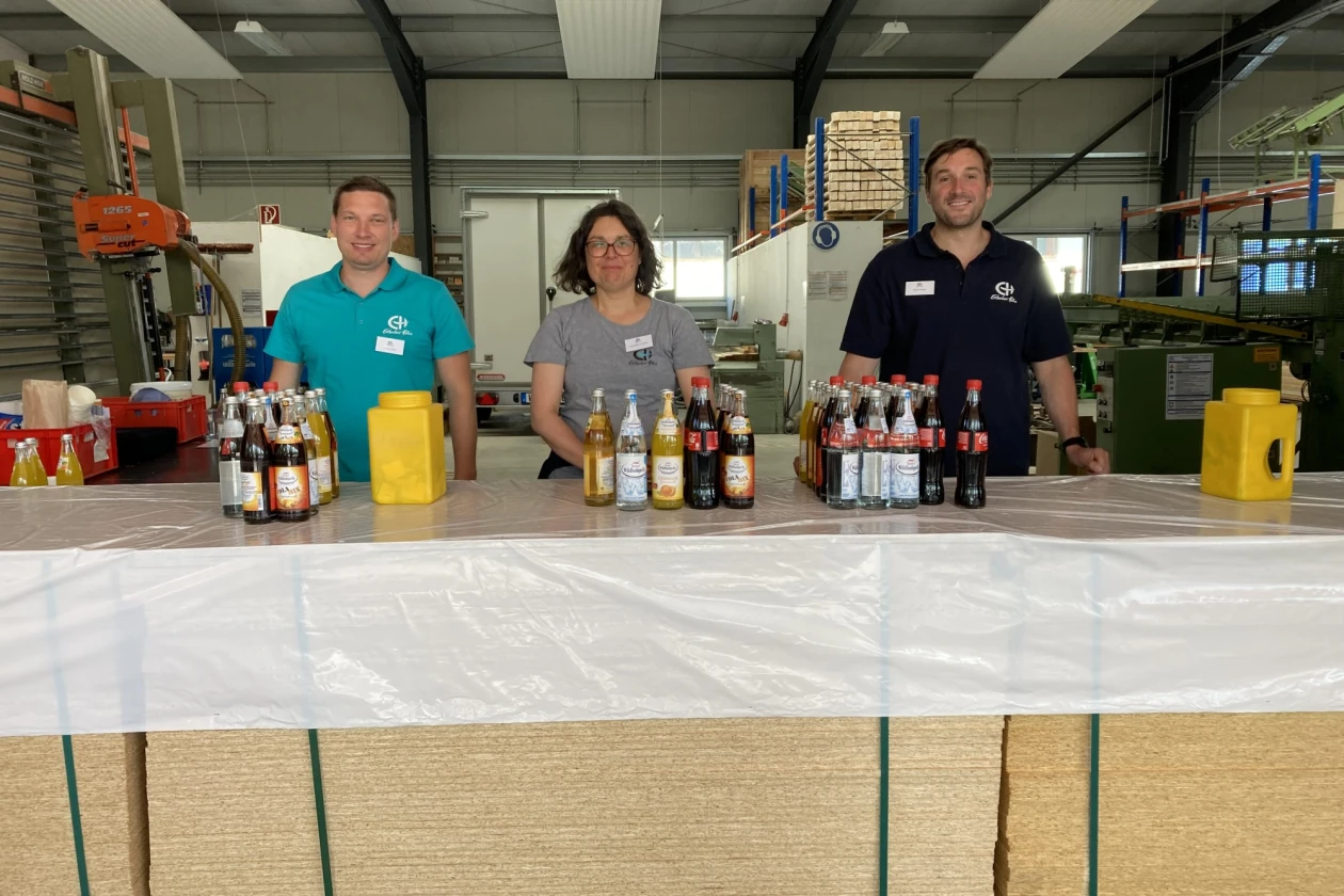 Drei Personen stehen hinter einer Theke, auf der verschiedene Getr&auml;nke stehen. Im Hintergrund ist eine Betriebshalle mit Maschinen und Regalen zu sehen.