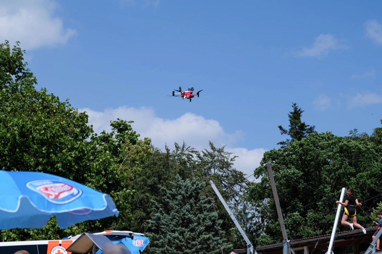 Eine Drohne fliegt im blauen Himmel &uuml;ber einem Hintergrund aus B&auml;umen.