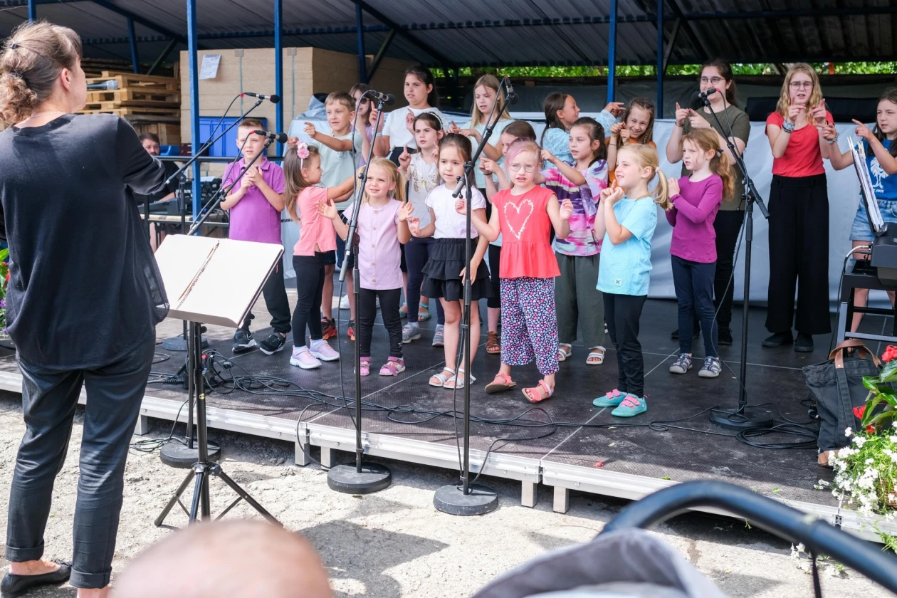 Ein Kinderchor steht auf einer B&uuml;hne. Im Vordergrund ist eine Frau an einem Notenst&auml;nder zu sehen.