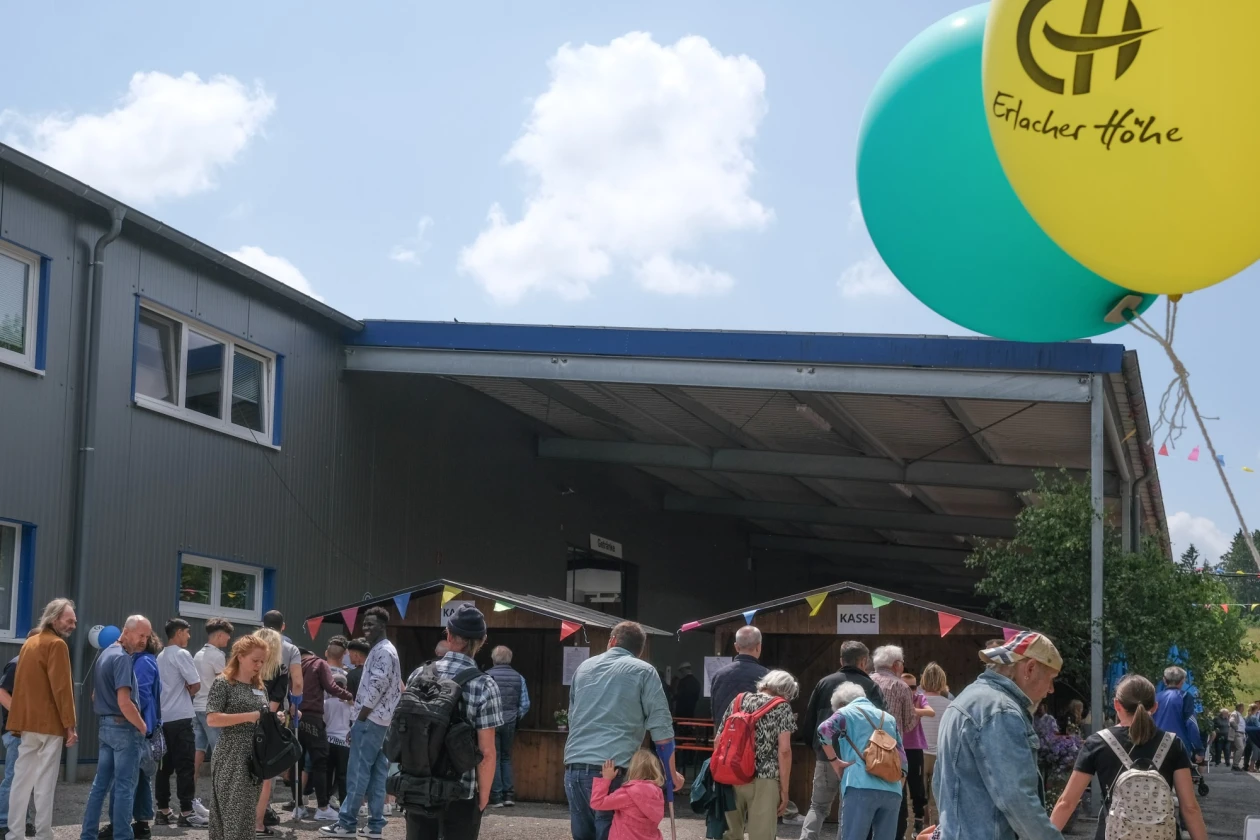 Menschen stehen im Festbetrieb vor zwei Kassenbuden. Im Hintergrund ist ein &uuml;berdachter Platz eines Firmengeb&auml;udes zu sehen. Im Vordergrund fliegen zwei Luftballons am Bildrand.