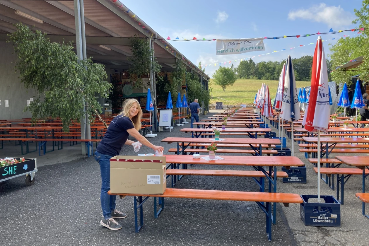 Leere Festb&auml;nke und geschlossene Sonnenschirme sind zu sehen. Auf einer Festbank steht ein ge&ouml;ffneter Karton, aus dem die Frau etwas herausholt und auf den Tisch legt.