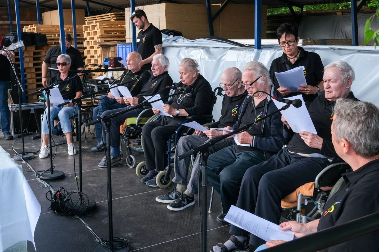 Eine Gruppe &auml;lterer Menschen, teilweise im Rollstuhl, in schwarzen Oberteilen sitzt vor Mikrofonen. Sie singen auf einer B&uuml;hne.