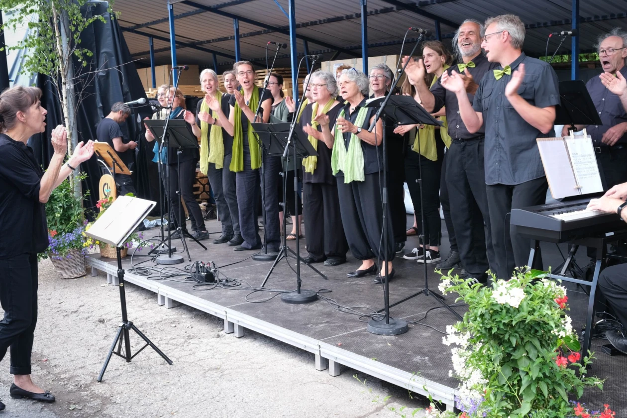 Ein Chor aus erwachsenen S&auml;ngerinnen und S&auml;ngern steht auf einer B&uuml;hne und wird von einer Frau an einem Notenst&auml;nder dirigiert. Ein Mann am E-Piano begleitet musikalisch.