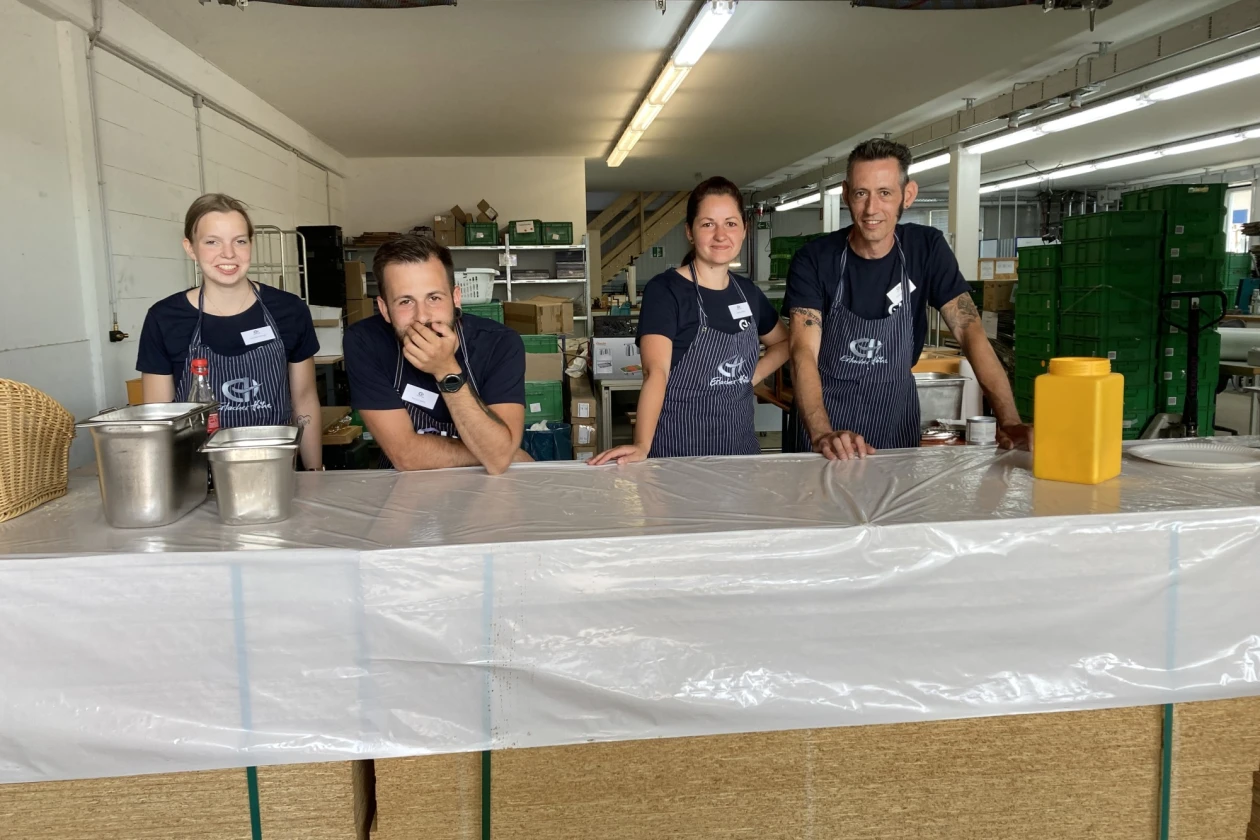 Vier Personen l&auml;cheln in die Kamera. Sie stehen hinter einer Theke,