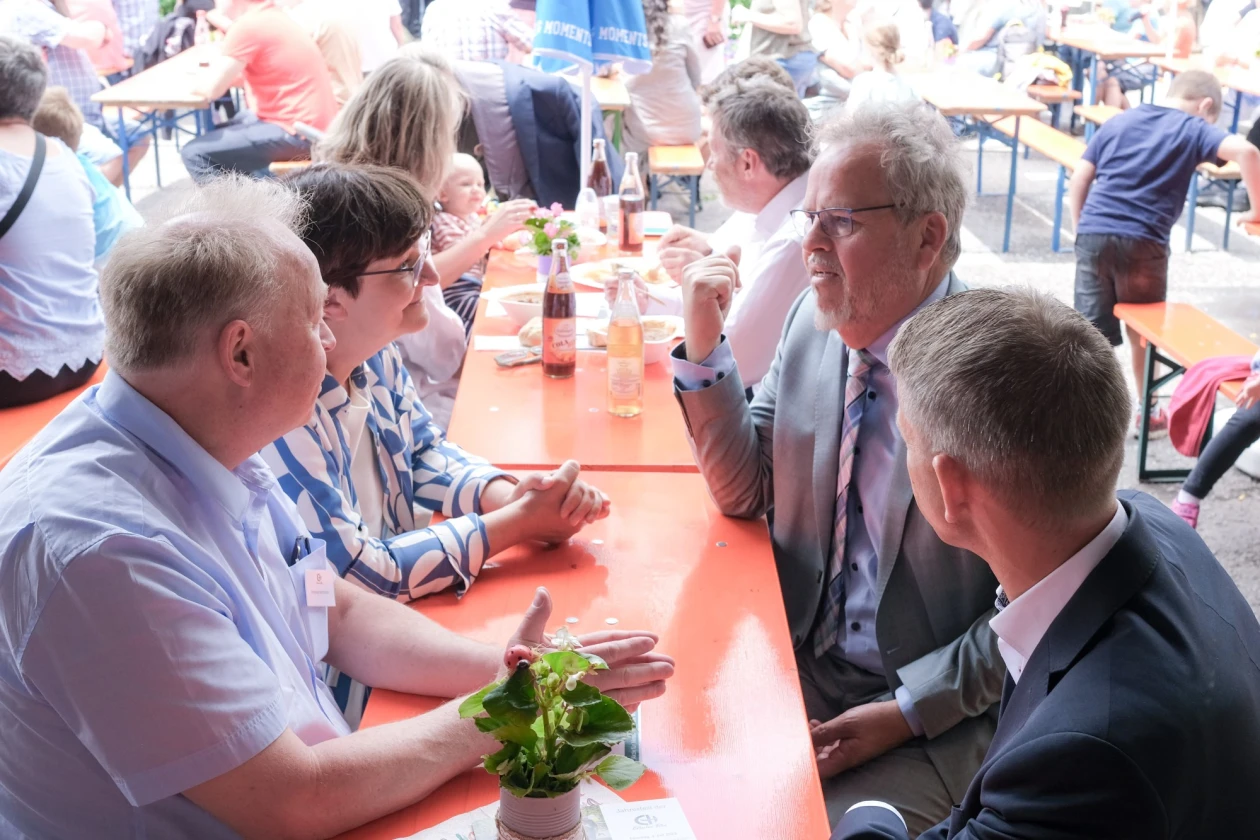 Mehrere Personen sitzen an Festgarnituren, genie&szlig;en Speisen und Getr&auml;nke und unterhalten sich.