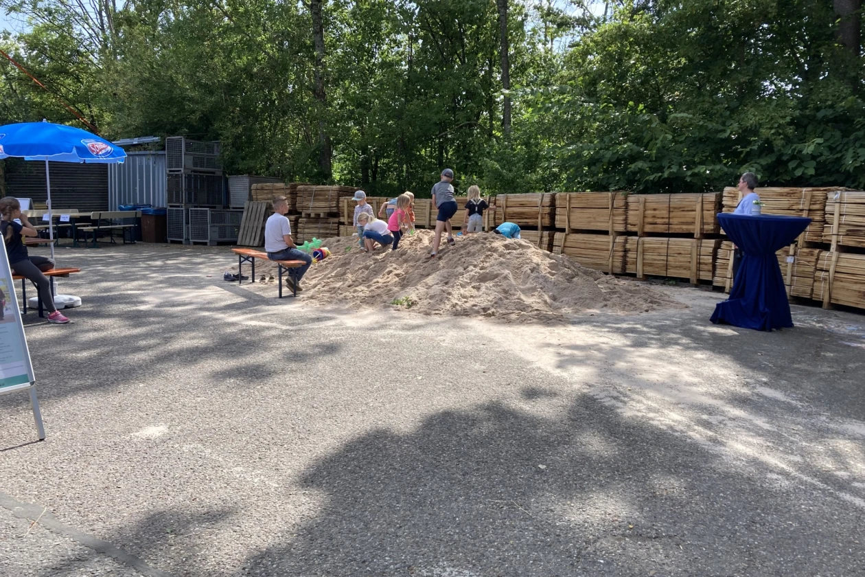 Zu sehen ist Sandberg, auf der Kinder spielen. Im Hintergrund sehen Holzlatten, ein ge&ouml;ffneter Sonnenschirm und B&auml;ume zu sehen.