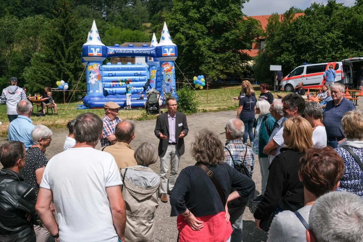 Eine Menschengruppe steht und h&ouml;rt einem Mann im Jackett zu. Im Hintergrund ist eine Kinderh&uuml;pfburg und ein Fahrzeug des Rettungsdienstes zu sehen.