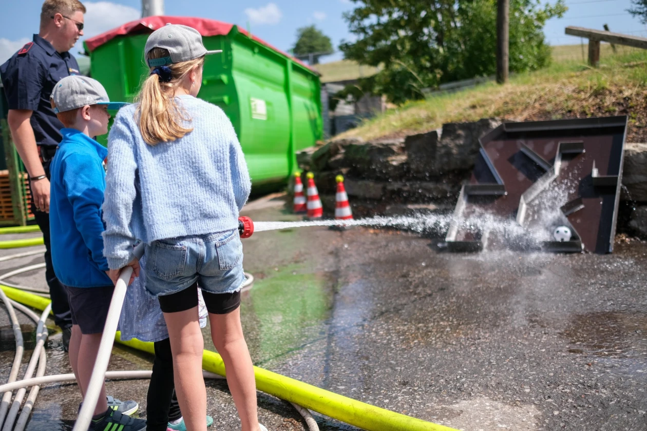 Zwei Kinder spritzen mit einem Feuerwehrschlauch. Im Hintergrund ist ein junger Feuerwehrmann zu sehen.