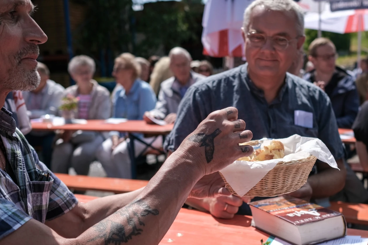 Zwei M&auml;nner sitzen im Vordergrund. Sie teilen sich Brot aus einem Korb. Im Hintergrund sitzen Menschen auf Festb&auml;nken