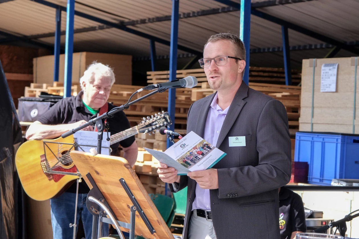 Ein &auml;lterer Mann mit einer akustischen Gitarre und ein Mann im grauen Anzug stehen vor Holzpaletten. Der Mann im Jackett steht am Rednerpult mit Mikrofon und h&auml;lt eine Brosch&uuml;re der Erlacher H&ouml;he in der Hand und redet ins Publikum.