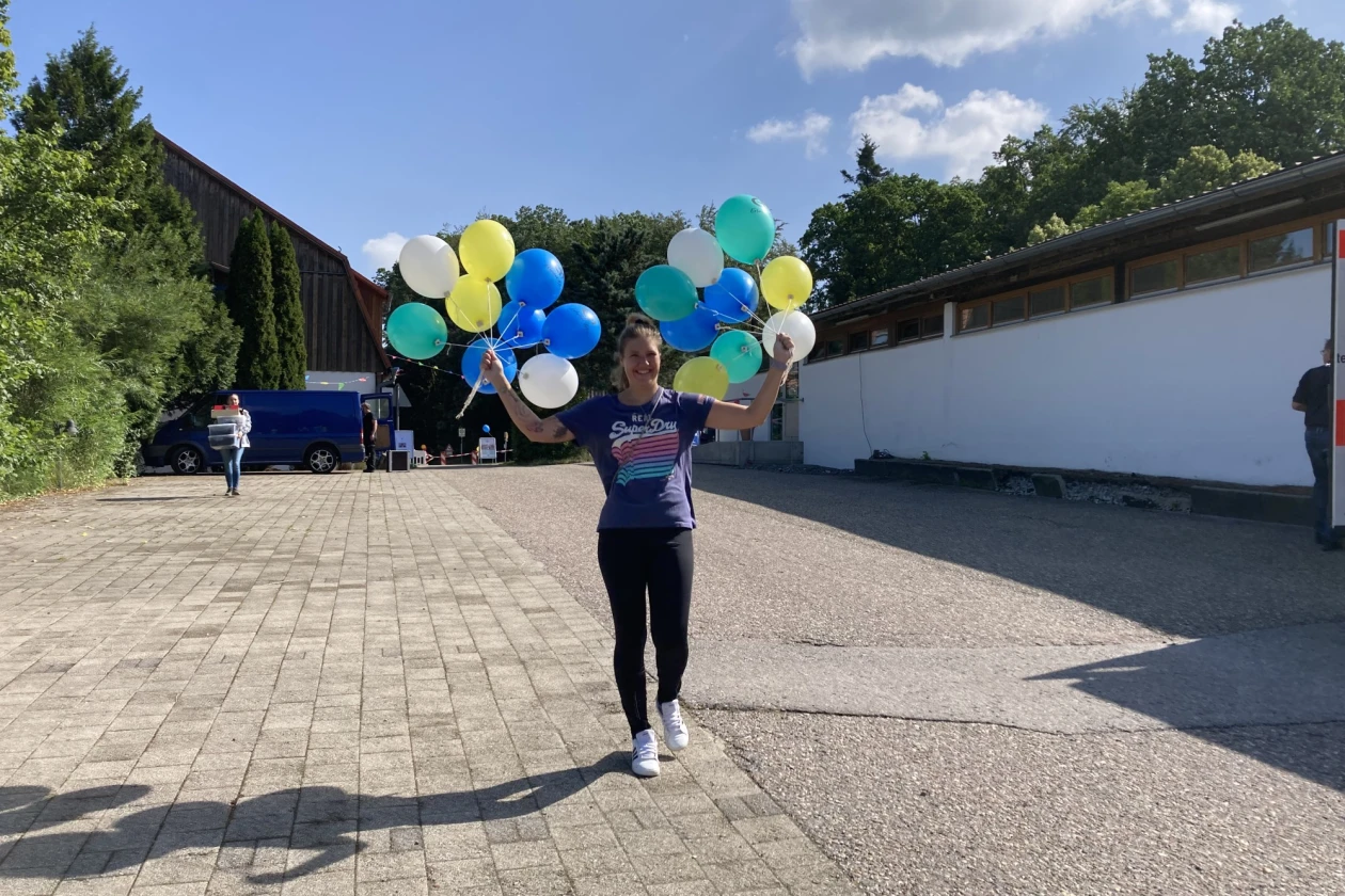 Eine Frau steht auf gepflastertem Boden und h&auml;lt in beiden H&auml;nden mehrere bunte Luftballons. Im Hintergrund sind eine Person und ein blauer Transporter zu sehen.