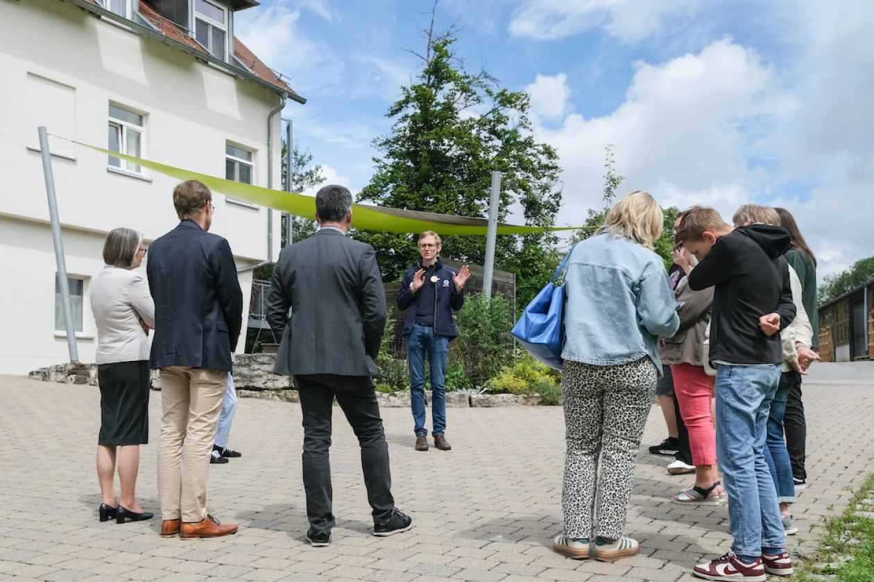 Eine Menschengruppe h&ouml;rt aufmerksam einem jungen Mann im Outfit mit dem Logo der Erlacher H&ouml;he zu. Im Hintergrund ist ein Wohngeb&auml;ude zu sehen.