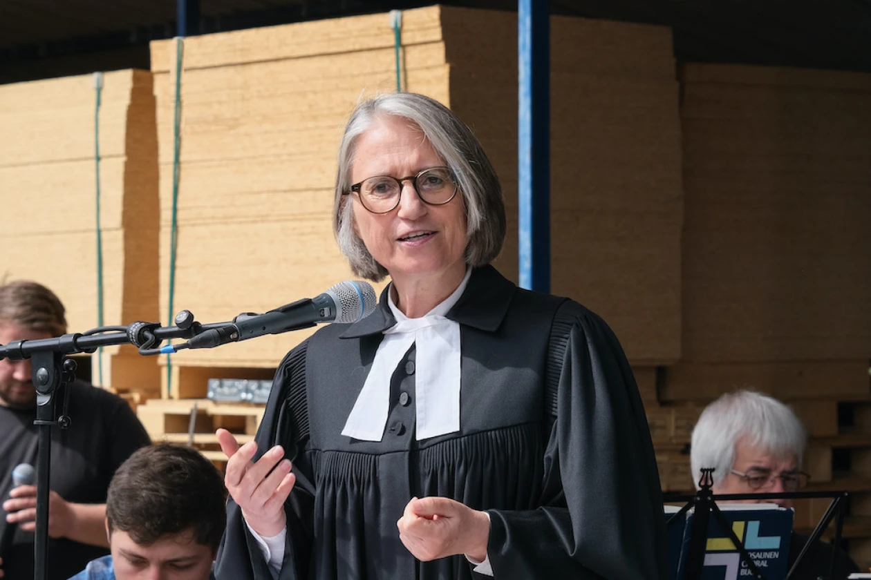 Eine grauhaarige Frau im Talar predigt auf einer B&uuml;hne im Freien.