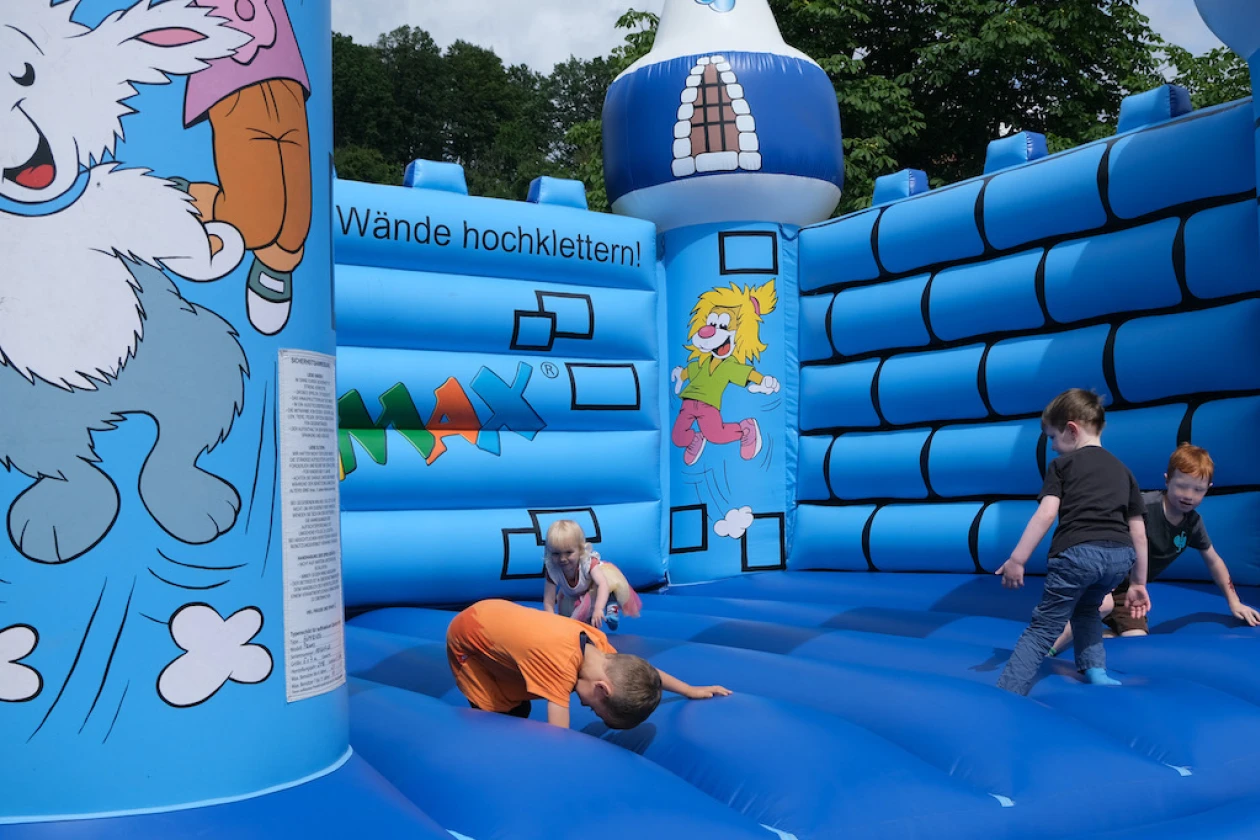 Eine blaue H&uuml;pfburg mit bunten Cartoon-Figuren, auf der mehrere Kinder herumklettern und spielen. Die Burg hat verschiedene Elemente wie W&auml;nde und S&auml;ulen und steht im Freien auf einem Rasengel&auml;nde mit B&auml;umen im Hintergrund.