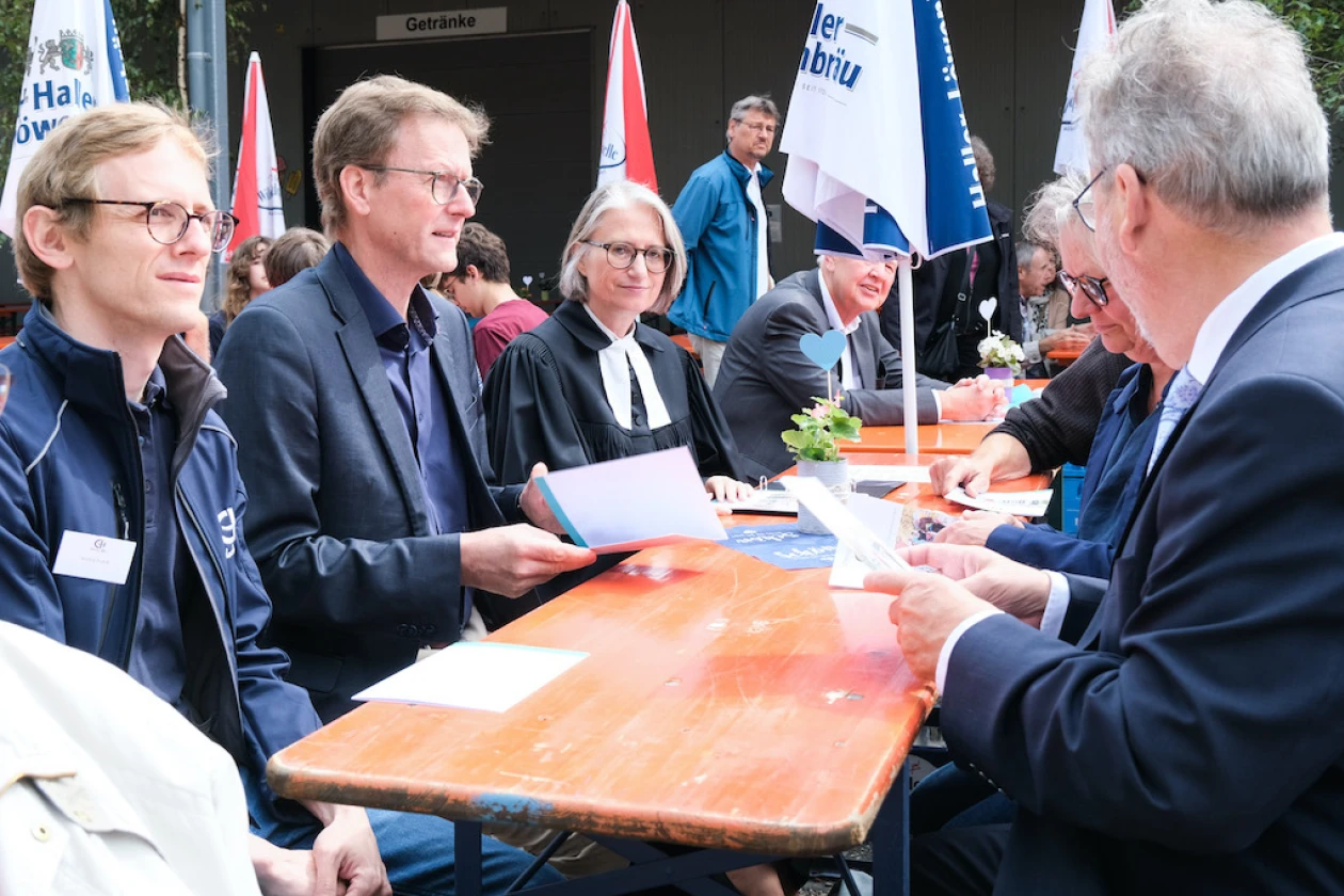 Mehrere Personen sitzen an einem Holztisch, offenbar bei einem Fest. Sie tragen verschiedene Kleidung wie Anz&uuml;ge und Jacken und scheinen in eine Diskussion vertieft zu sein.