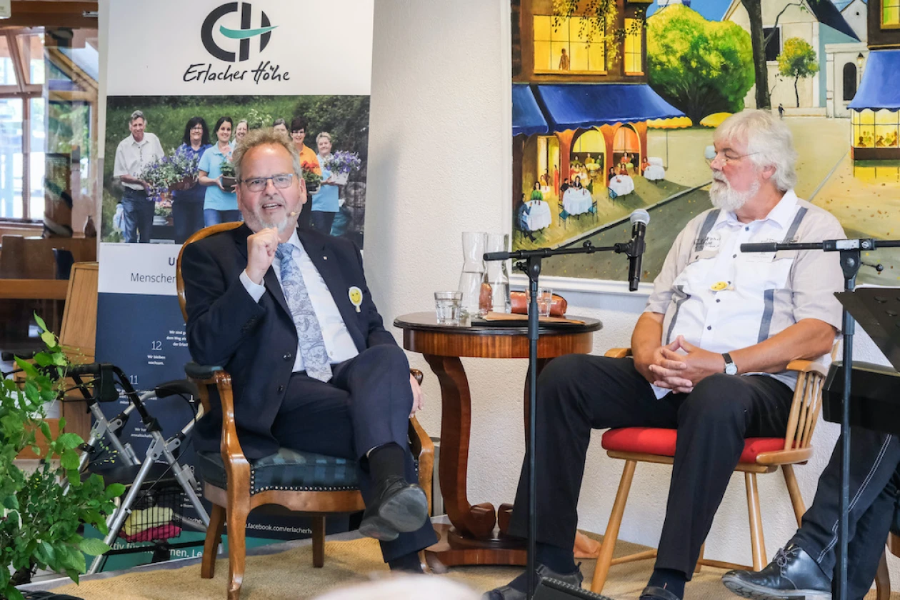 Zwei M&auml;nner sitzen in Sesseln bei einer Veranstaltung, einer im dunklen Anzug mit Brille und einer im wei&szlig;en Hemd mit wei&szlig;em Bart. Im Hintergrund ein Poster und farbenfrohe Wandkunst, die eine lockere und offene Gespr&auml;chssituation zeigt.