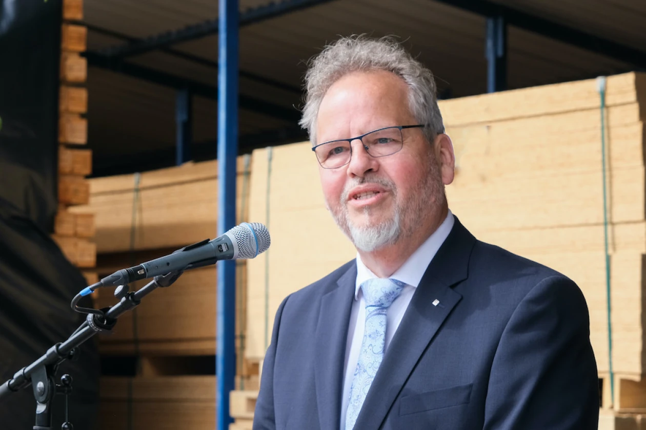 Ein grauhaariger Mann mit Bart und Brille steht vor einem Mikrofon und spricht. Im Hintergrund sind gelagerte Holzplatten zu sehen.