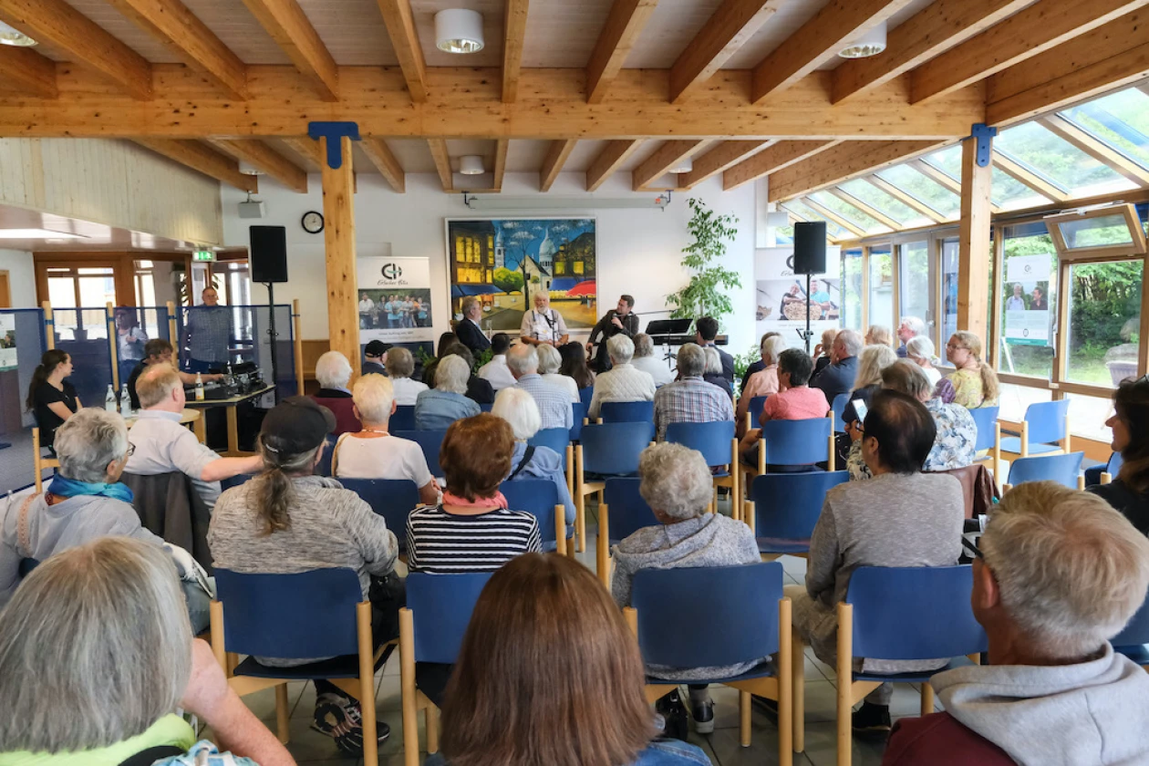 Mehrere Menschen sitzen in einem hellen, holzbalkengedeckten Raum mit gro&szlig;en Fenstern und h&ouml;ren einem Vortrag zu. Blaue St&uuml;hle sind in Reihen aufgestellt, und im Hintergrund ist ein farbenfrohes Kunstwerk zu sehen.