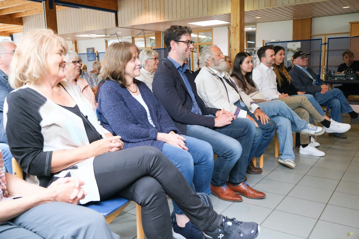 Viele unterschiedliche Menschen sitzen in Reihen in einem Zuh&ouml;rerraum. Sie l&auml;cheln und h&ouml;ren aufmerksam zu.