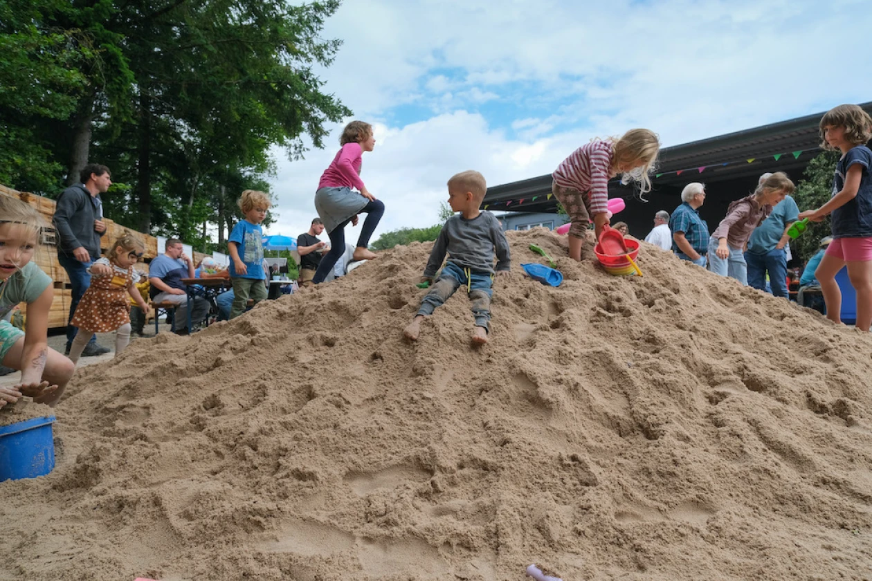 Mehrere Kinder spielen und klettern auf einem gro&szlig;en Sandhaufen, w&auml;hrend Erwachsene im Hintergrund zusehen. Die Szene findet im Freien statt, mit B&auml;umen und blauem Himmel, und die Kinder scheinen Spa&szlig; beim Erkunden und Spielen im Sand zu haben.