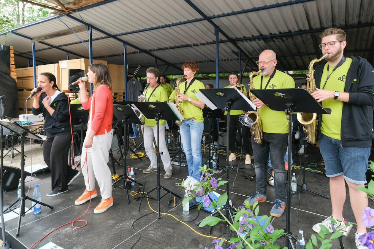 Mehrere Personen stehen auf einer B&uuml;hne. Zwei Frauen haben Mikrofone in der Hand und singen, w&auml;hrend die anderen mit verschiedenen Instrumenten dahinter stehen. Die Personen im Hintergrund tragen einheitlich gr&uuml;ne T-Shirts.