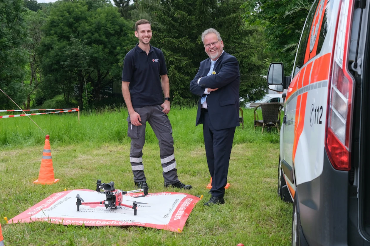 Zwei M&auml;nner stehen neben einem Fahrzeug des Deutschen Roten Kreuzes und blicken auf eine Drohne, die auf einer roten Matte, umgeben von gr&uuml;ner Wiese und B&auml;umen steht. Die M&auml;nner tragen einen dunklen Arbeitsanzug und einen blauen Anzug und l&auml;cheln in die Kamera.
