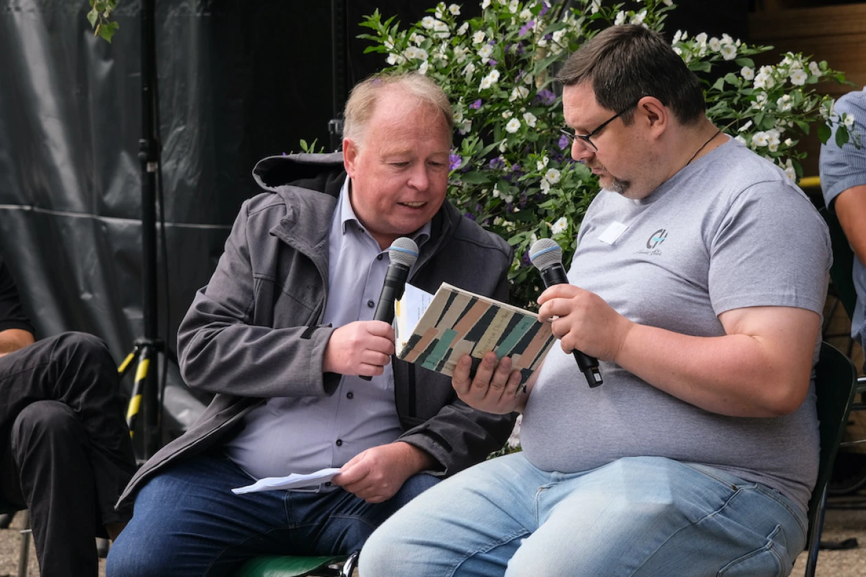 Zwei M&auml;nner sitzen nebeneinander mit Mikrofonen in der Hand. Sie sprechen Texte, die sie aus einem Notizbuch lesen.