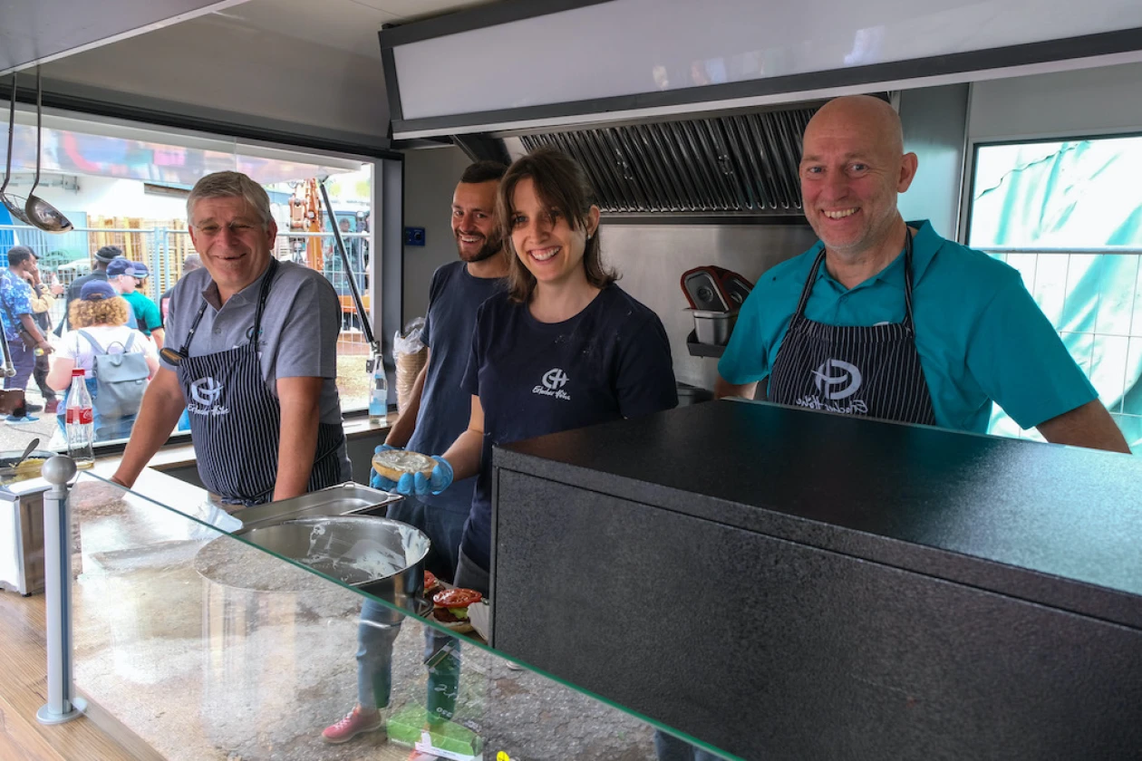 Drei M&auml;nner und eine Frau Sch&uuml;rzen mit dem Logo der Erlacher H&ouml;he stehen in einem Foodtruck und l&auml;cheln freundlich in die Kamera, bereit f&uuml;r neuen Service.