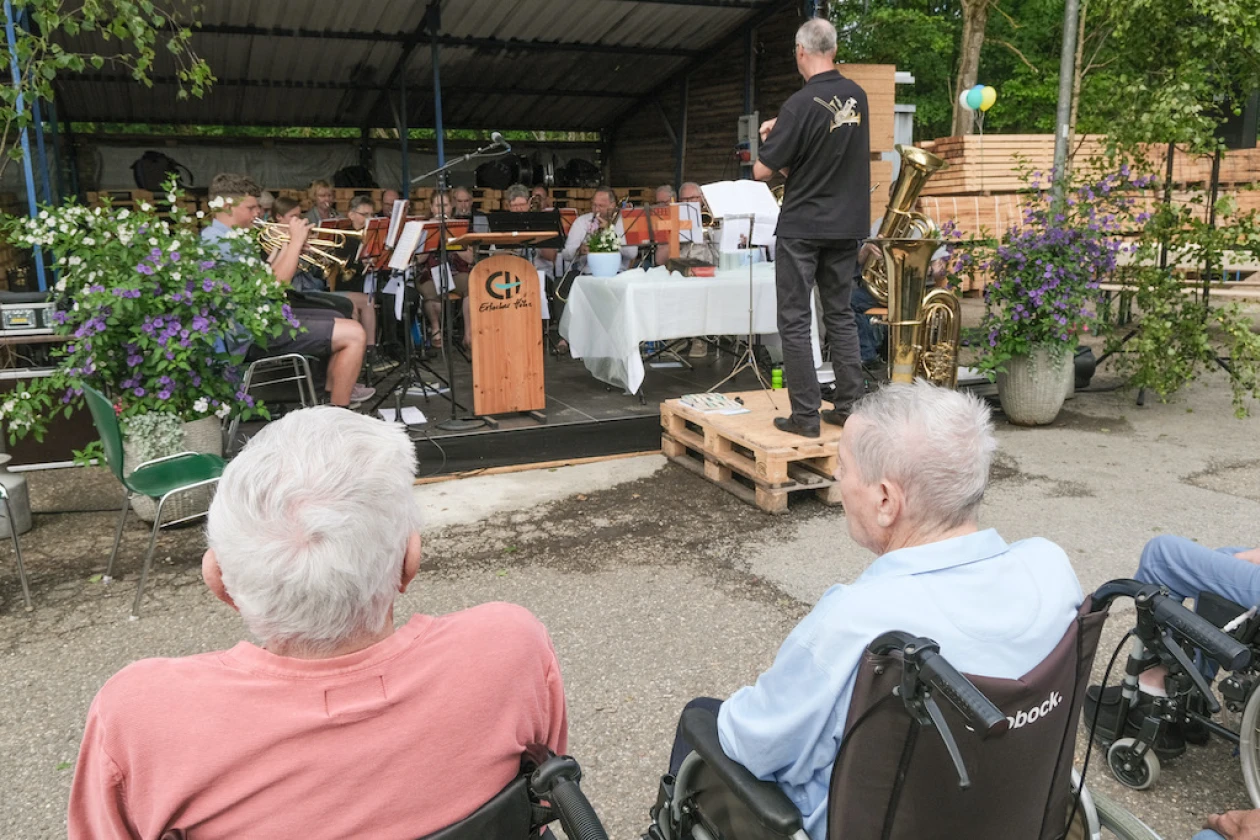 Auf einer B&uuml;hne sitzen Musiker mit Blechblasinstrumenten, die von einem schwarz gekleideten Mann dirigiert werden. Im Vordergrund sitzen Menschen im Rollstuhl und h&ouml;ren zu.