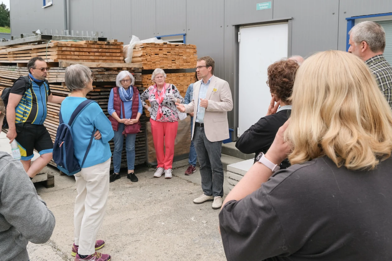 Eine Gruppe Menschen steht vor einer Industriehalle und h&ouml;rt aufmerksam einem Mann in hellem Hemd und Jackett zu. Im Hintergrund sind lagernde Holzstapel zu sehen.