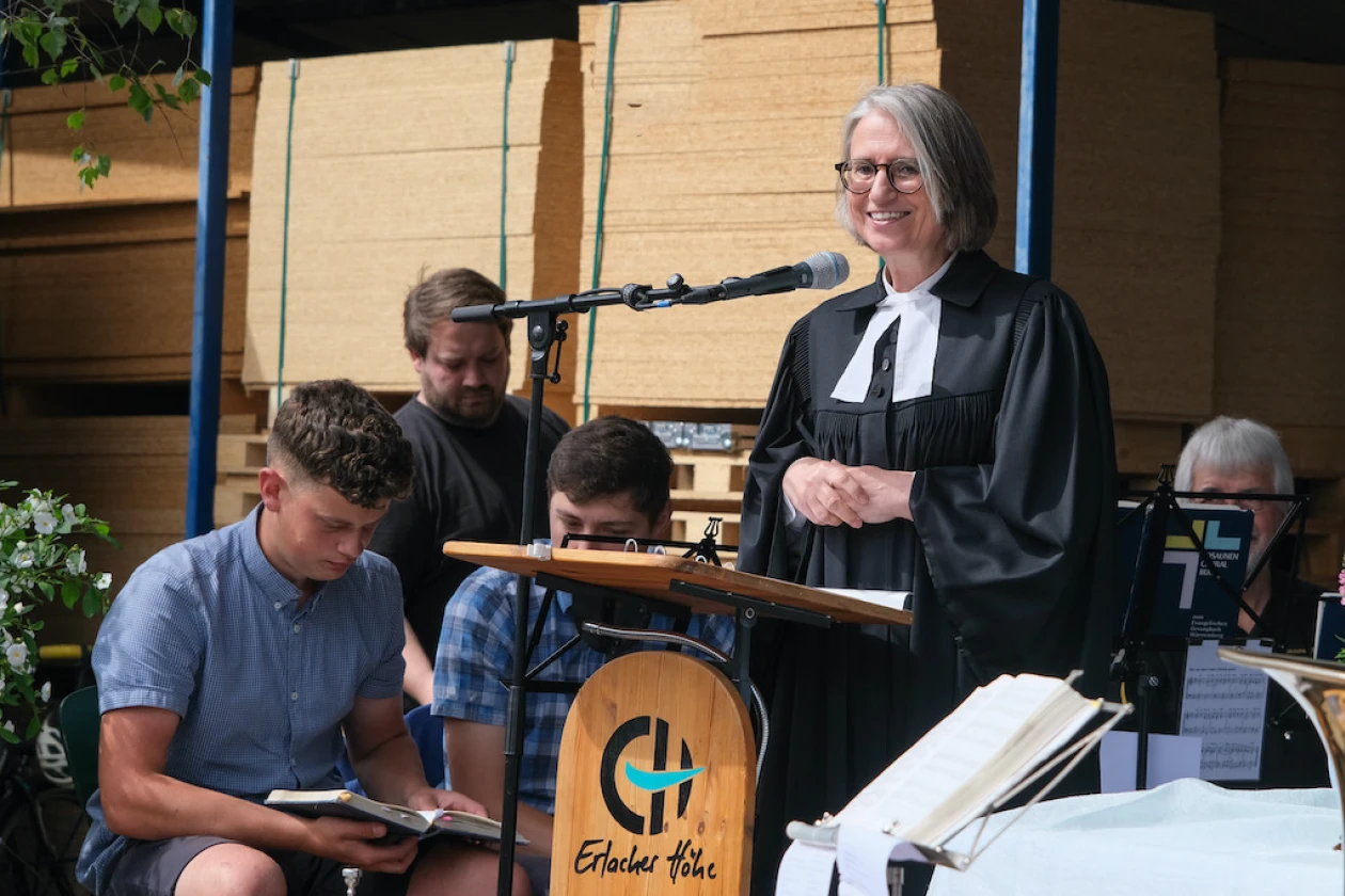 Eine grauhaarige Frau im Talar predigt auf einer B&uuml;hne im Freien. Sie hat ihre H&auml;nde vor dem K&ouml;rper ineinandergelegt, spricht und l&auml;chelt in die Zuschauermenge.