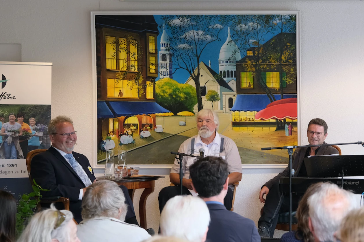Drei M&auml;nner sitzen auf einer B&uuml;hne. Der &auml;ltere Mann mit wei&szlig;em Haar h&auml;lt einen Vortrag vor einem Publikum, w&auml;hrend im Hintergrund ein Gem&auml;lde einer abendlichen Stadtszene mit Kirche und beleuchteten Geb&auml;uden zu sehen ist.