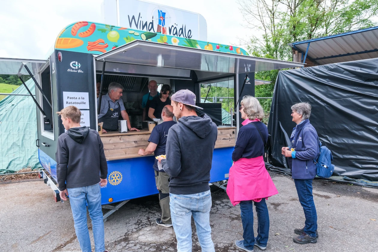 Mehrere Personen stehen an einem hellblauen Verkaufsanh&auml;nger namens "Windr&auml;dle" und warten auf Essen.