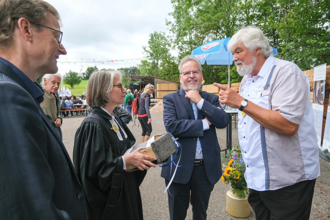 Eine Gruppe von vier Menschen unterh&auml;lt sich bei einer Veranstaltung im Freien, mit B&auml;umen und Festbetrieb im Hintergrund. Verschiedene Personen in unterschiedlicher Kleidung, von Anzug &uuml;ber Talar bis Freizeitkleidung, stehen in einer lockeren Gespr&auml;chsrunde zusammen.