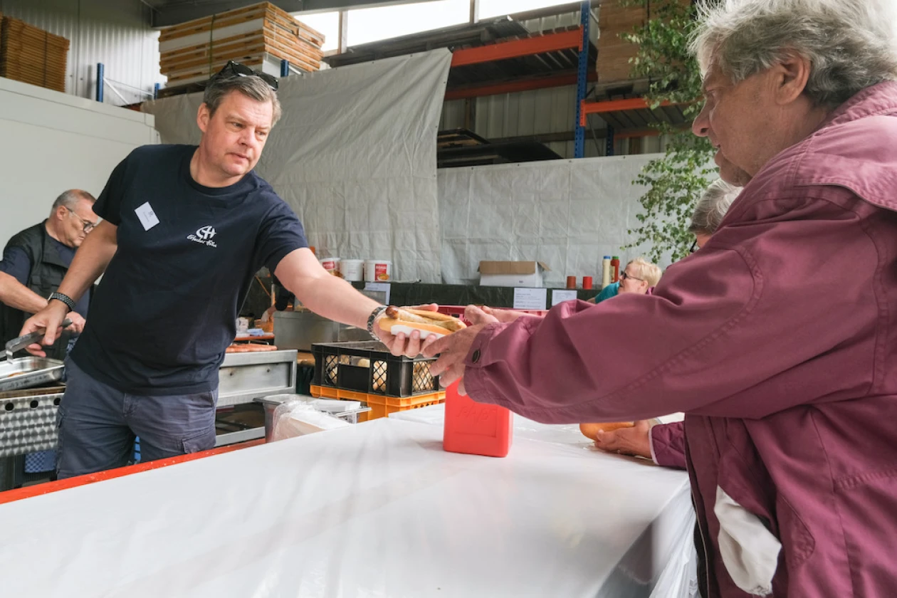 Ein Mann in schwarzem T-Shirt &uuml;berreicht einem &auml;lteren Mann in hellroter Jacke eine Bratwurst im Br&ouml;tchen &uuml;ber die Theke.