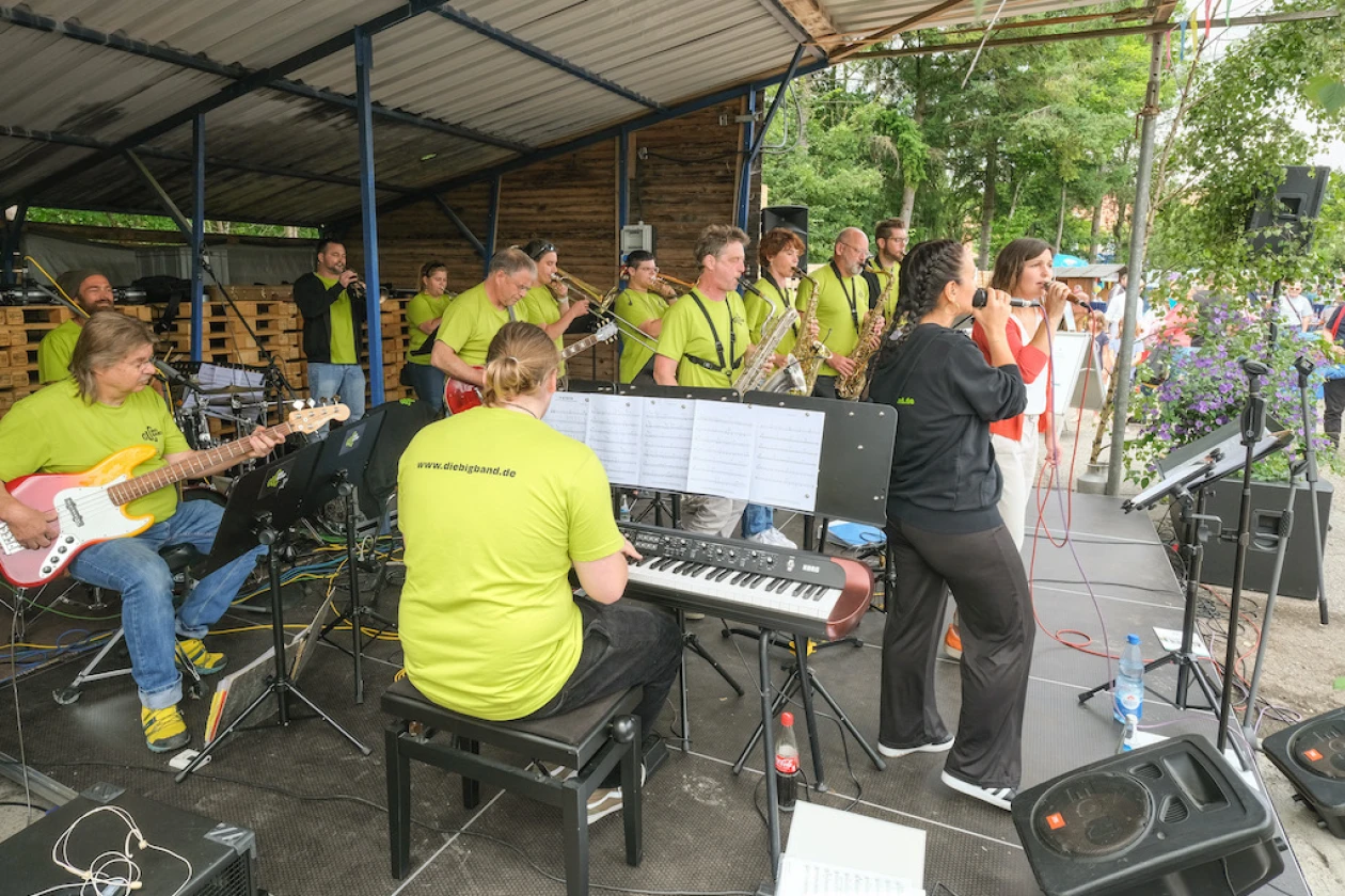 Eine Band spielt auf einer &uuml;berdachten B&uuml;hne mit Keyboard, E-Gitarre und mehreren Musikern in gelben Shirts und zwei S&auml;ngerinnen im Vordergrund.