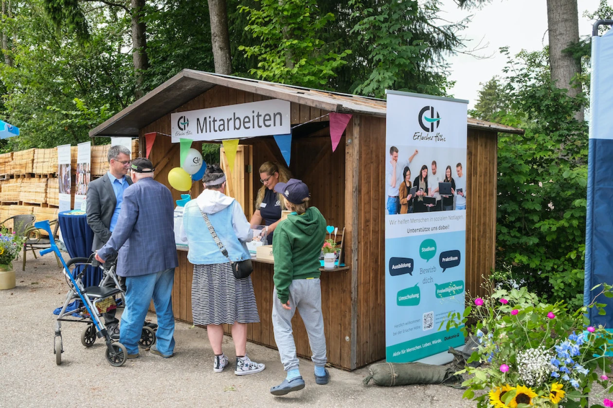 Informationsstand mit Thema "Mitarbeiten" und einem Aufsteller. Mehrere Personen verschiedenen Alters stehen an und tauschen sich aus, umgeben von B&auml;umen und Blumen an einem sonnigen Tag.