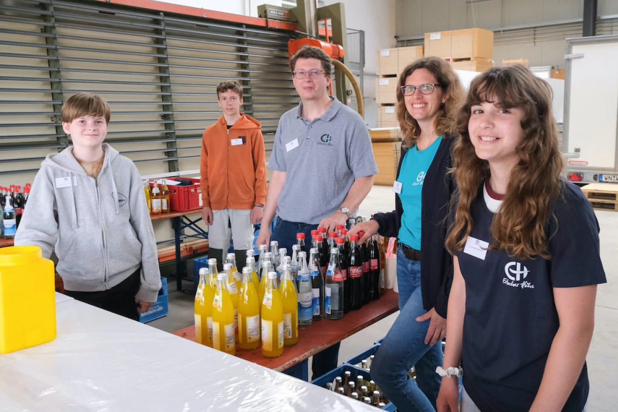 F&uuml;nf Personen stehen hinter einer Getr&auml;nketheke und blicken freundlich in die Kamera. Auf einem Holztisch stehen verschiedene Getr&auml;nke, unter dem Tisch weitere Getr&auml;nkekisten.