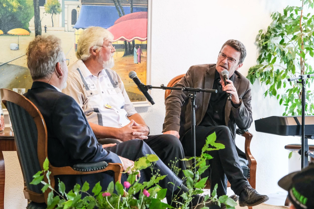 3 M&auml;nner sitzen auf einer B&uuml;hne mit Mikrofon. Der wei&szlig;haarige Mann moderiert die Beitr&auml;ge der anderen beiden M&auml;nner.