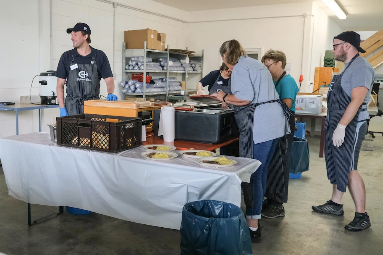 Mehrere Personen mit Sch&uuml;rzen bekleidet stehen in einer Halle um eine Verpflegungsstation und portionieren Mahlzeiten. Im Hintergrund sind Regale zu sehen.